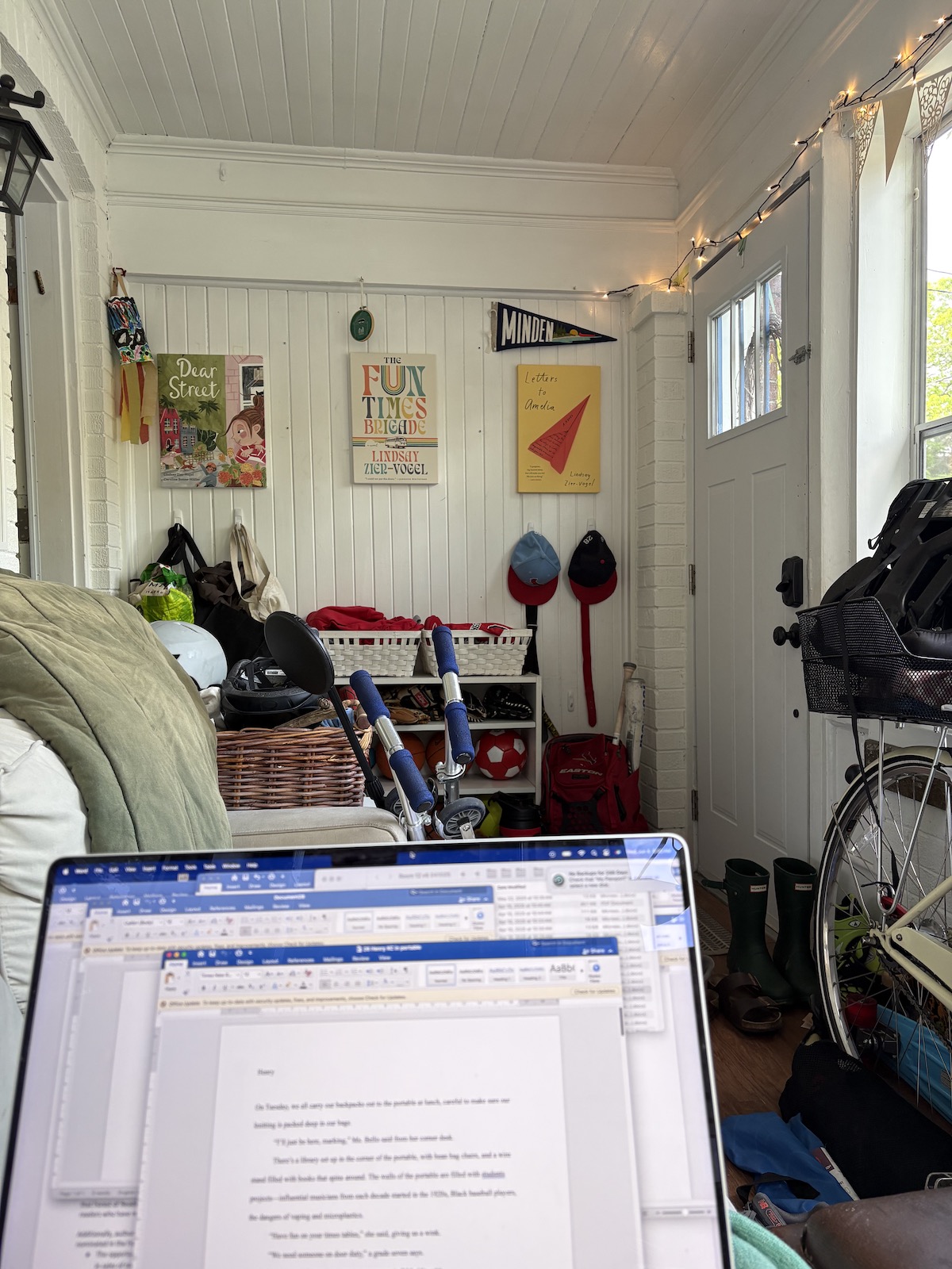 The view from Lindsay Zier-Vogel's writing spot. Her laptop screen is in the foreground, and in the background you can see her front room, the front door, the wheel of a bicycle, and fairy lights strung along the ceiling. Posters of her three book covers Dear Street, The Fun Times Brigade, and Letters to Amelia, are on the opposite wall.