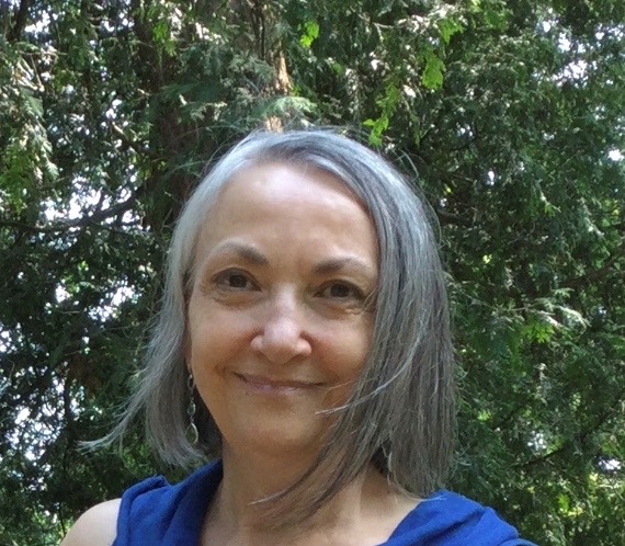 A photo of writer Karen Smythe. She is a light skin-toned woman with chin-length grey hair, wearing a blue sleeveless shirt and standing before a giant, leafy tree. She smiles slightly.
