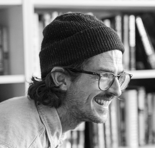 A black and white photo of writer James Cairns. He is a light skin toned man with longer dark hair that curls at the ends, sticking out from under a toque. He wears horn-rimmed glasses and stubble, and is laughing, almost in profile. He is pictured in front of a bookcase.