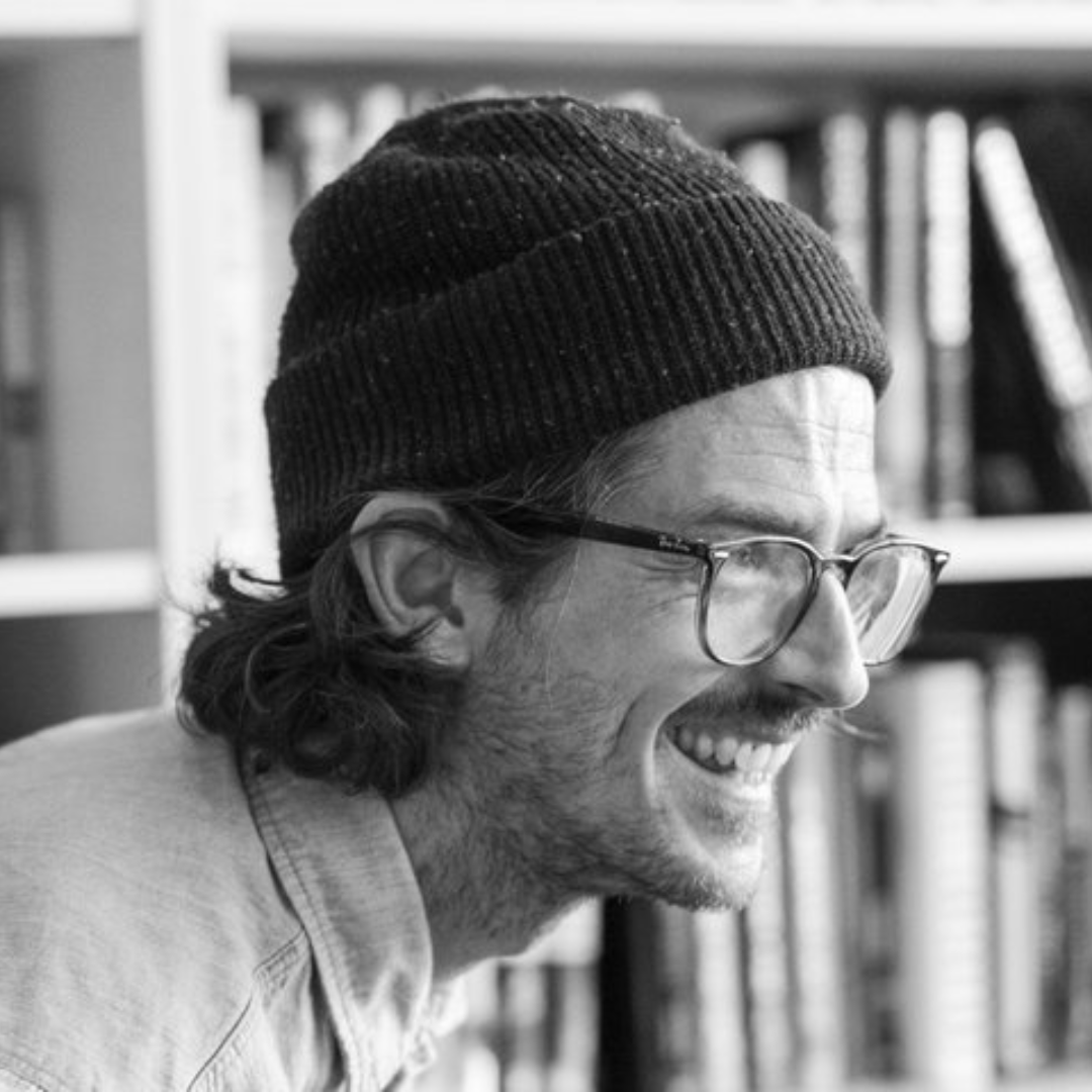 A black and white photo of writer James Cairns. He is a light skin toned man with longer dark hair that curls at the ends, sticking out from under a toque. He wears horn-rimmed glasses and stubble, and is laughing, almost in profile. He is pictured in front of a bookcase.