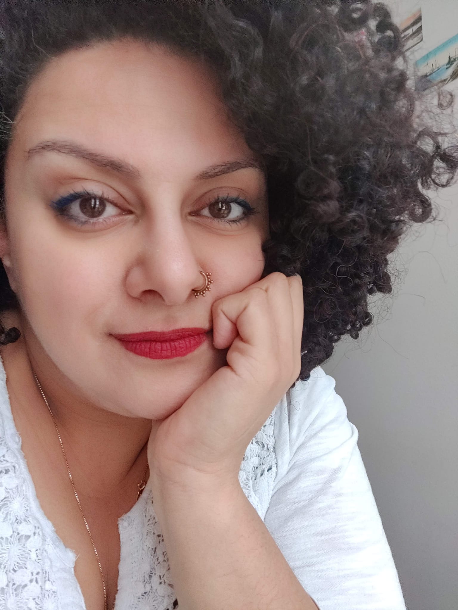 A photo of poet Carolyn Ramzy. She is a medium-skin toned Egyptian woman with very curly dark hair that is piled on and around her head, wearing red lipstick and perfectly-applied eyeliner. She rests her chin in her palm and stares into the camera.