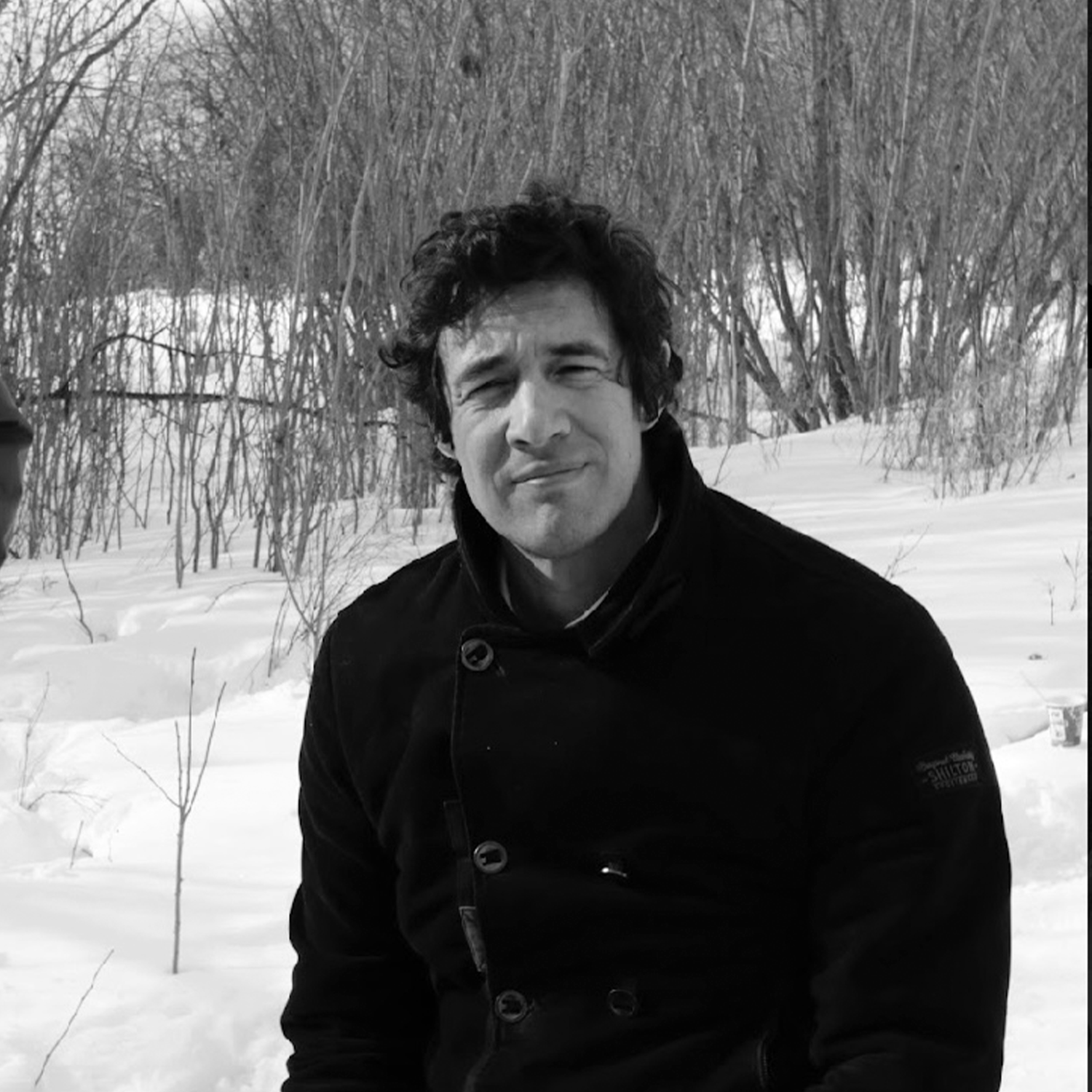 A black-and-white photo of author Sarain Frank Soonias. He is a Cree/Ojibwe writer with dark curly hair. He is standing outdoors in a snowy field. 