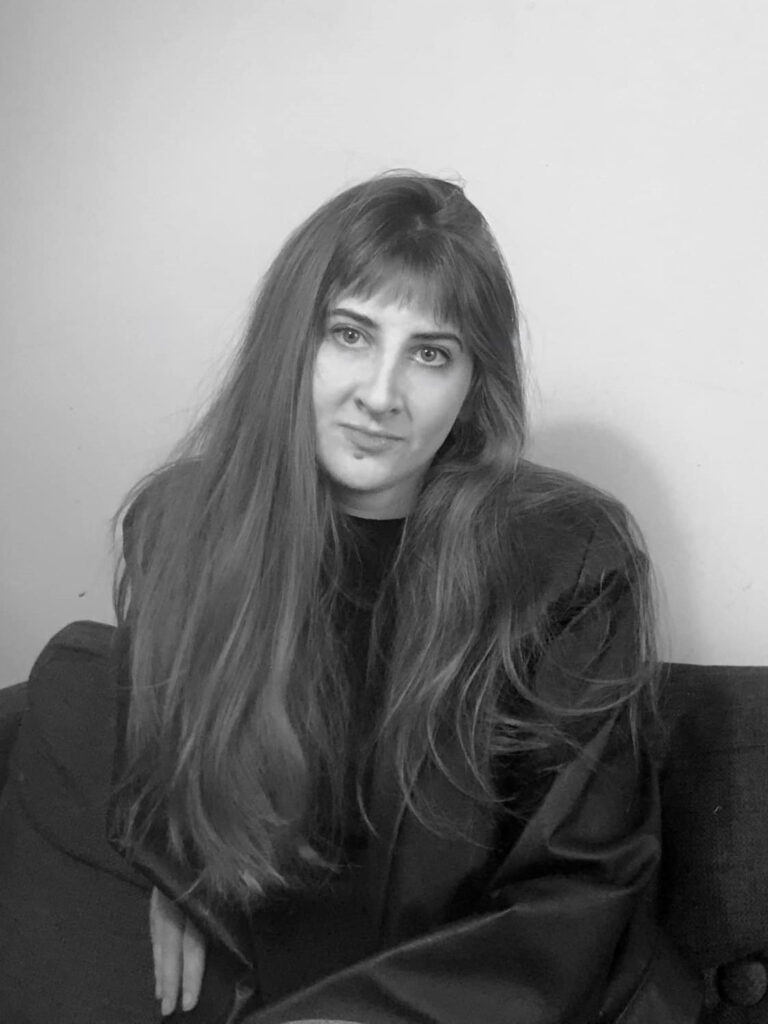 A black-and-white photo of author Miranda Schreiber. She is a light-skin-toned woman with long hair and bangs. She is wearing an oversized shirt and sitting on a couch.