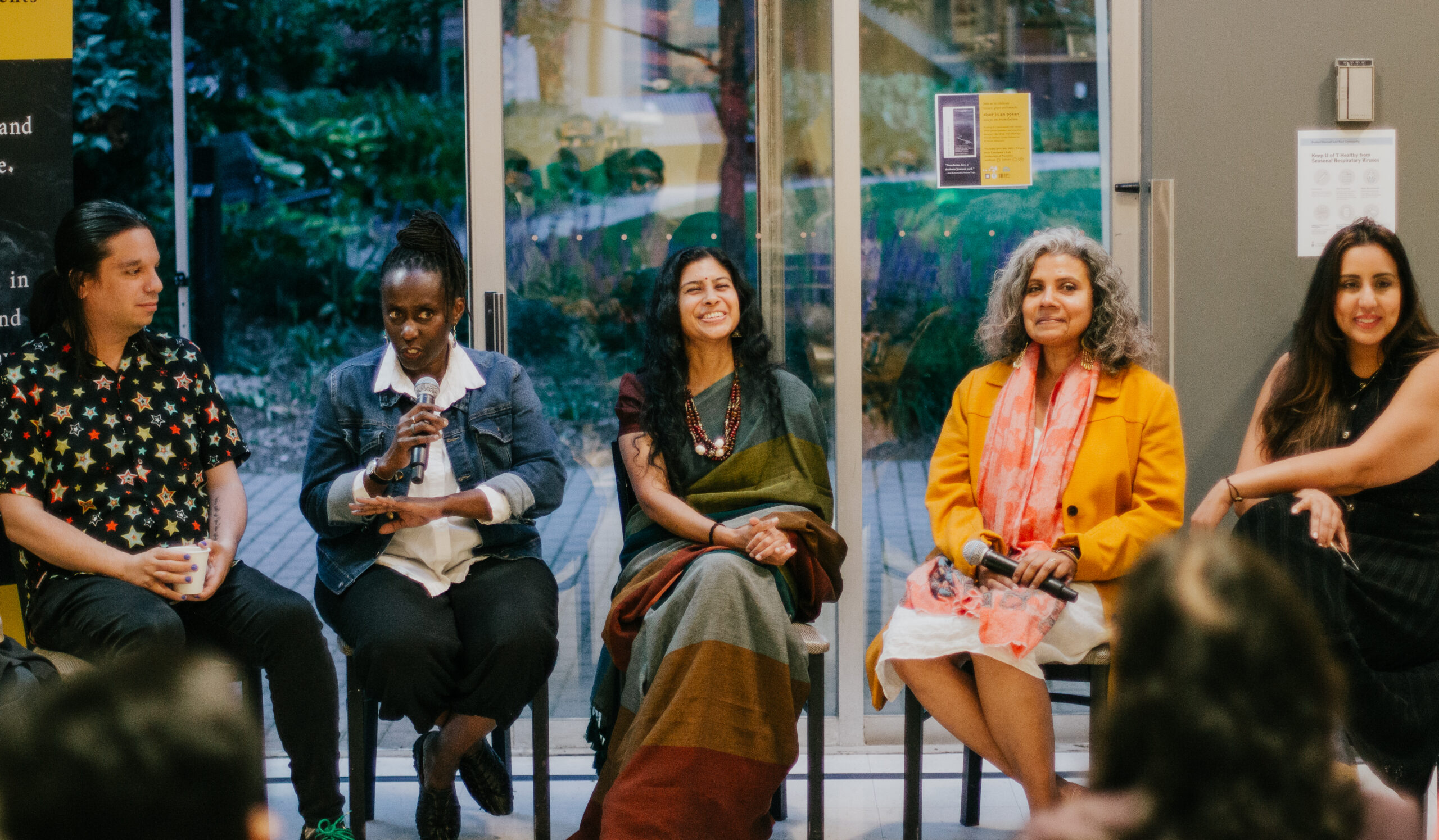 A photo of five authors and translators sitting on chairs in front of an audience. One of the authors is talking into a microphone while the others look out into the audience.
