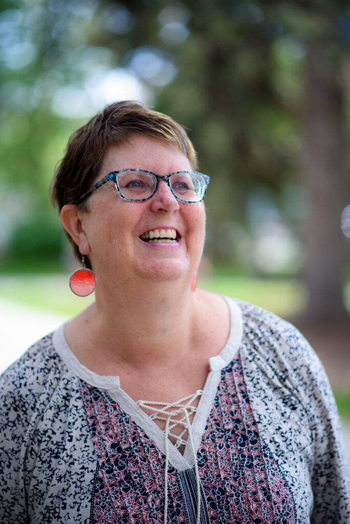A photo of Elizabeth J. Haynes. She is a light-skin-toned woman with short brown hair. She is wearing a multi-coloured shirt, red earrings, and blue-rimmed glasses. She is standing outdoors and laughing.