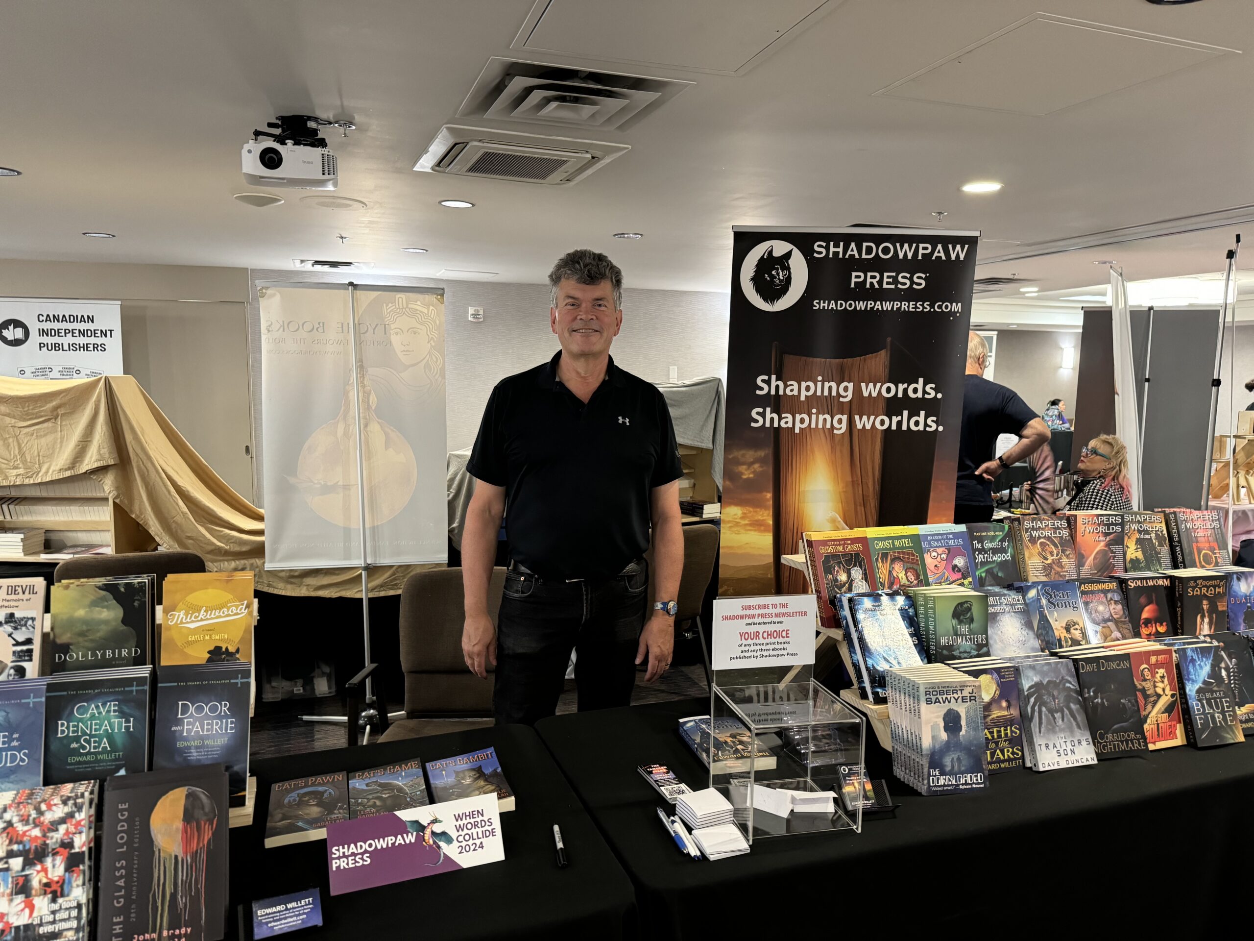 A photo of Edward Willett of Shadowpaw Press. He is a light-skin-toned man with short dark hair, wearing black jeans and a black shirt. He is standing at a booth and smiling into a camera. There are books behind him and a banner that reads Shadowpaw Press.