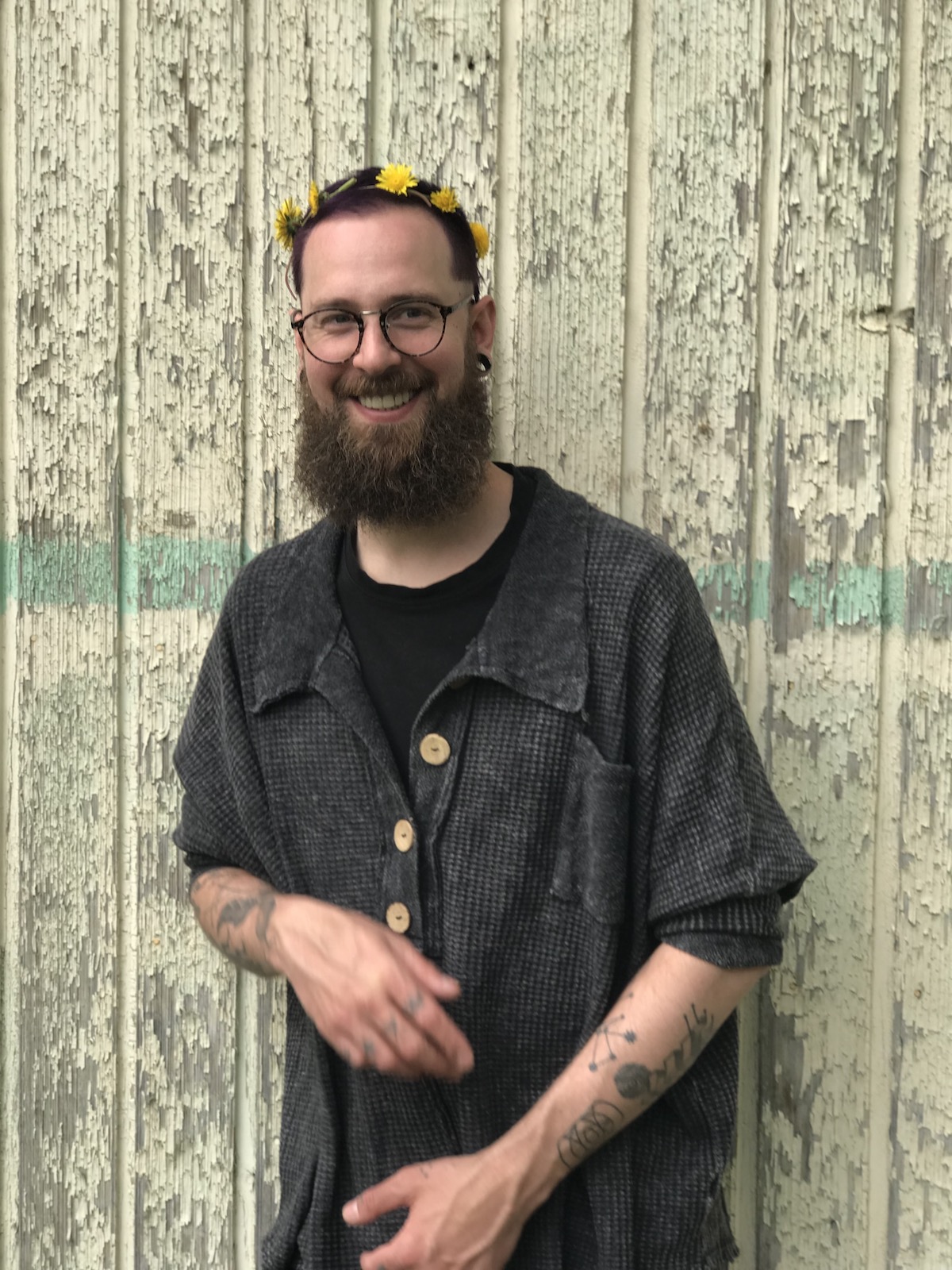 A photo of poet Kyle Flemmer. He is a tall, light skin-toned man with close-cropped dark hair and a bushy, dark beard. He wears glasses and a dark grey shirt, and is crowned with a string of dandelions. He looks to be mid laugh, his hands rising from his sides.