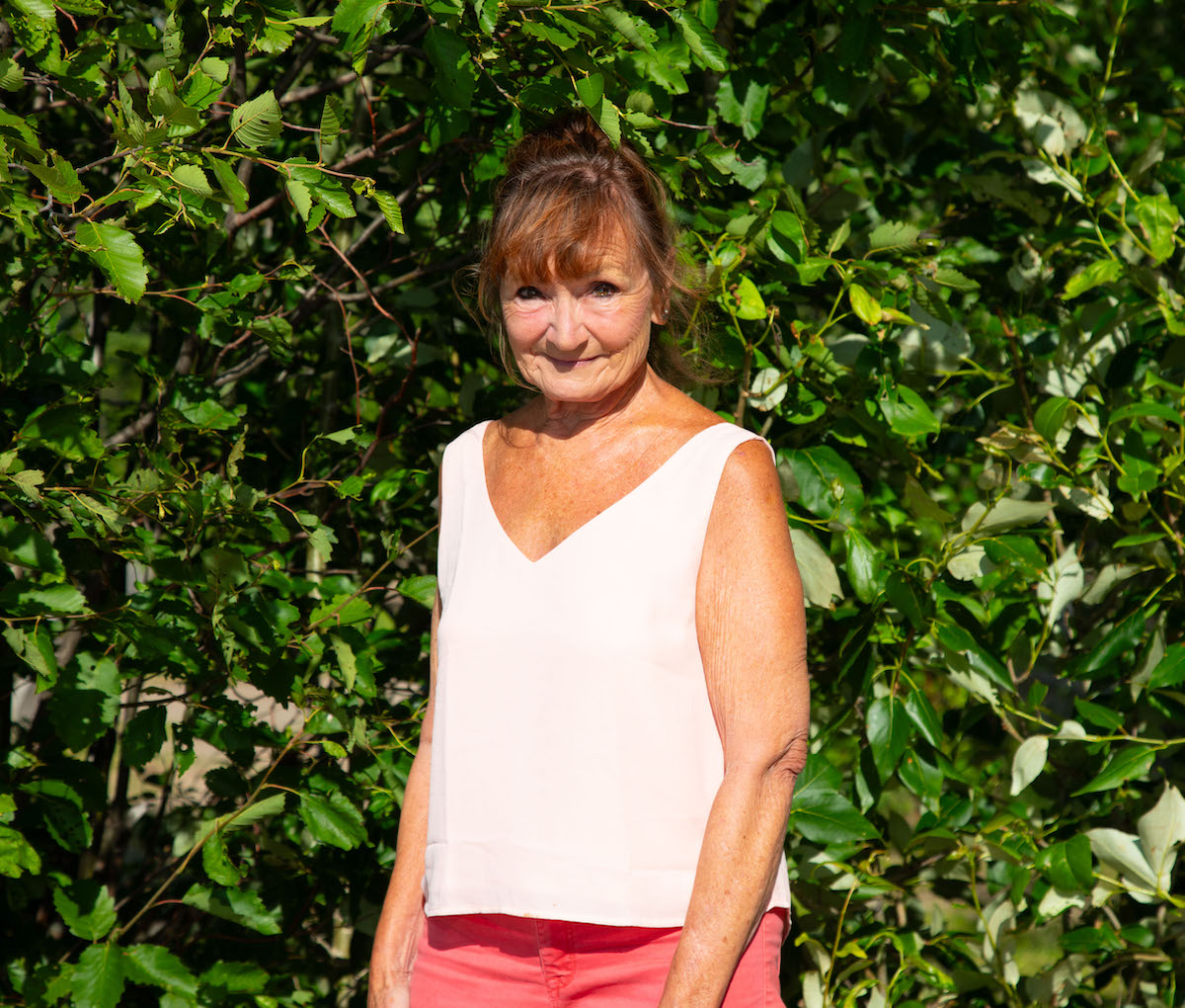 A photo of poet Lynda Monahan. She is a light skin toned woman with a golden tan, with reddish-brown hair cut into bangs and tied back into a ponytail, wearing a white sleeveless shirt and bright pink shorts. She stands in front of a leafy hedge, in the sunlight.