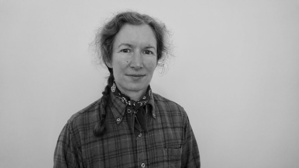 A black and white photo of Natalie Porter. She is a light skin-toned woman with medium coloured-hair worn in a braid, wearing a plaid shirt and bandana around her neck.