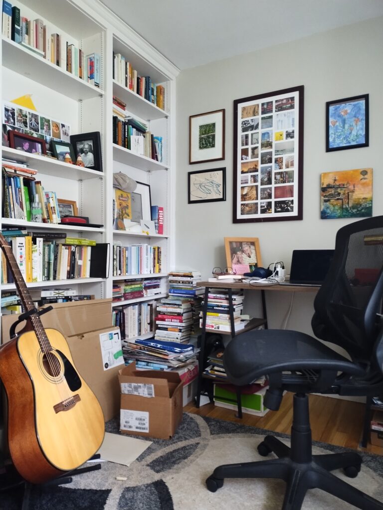 A photo of Nick Thran's office. There  are white bookshelves with books and framed photos on it, an acoustic guitar leaning against it, and a desk with an office chair on the opposite side. There are stacks of books on the floor beside the desk, and framed artwork on the wall. 