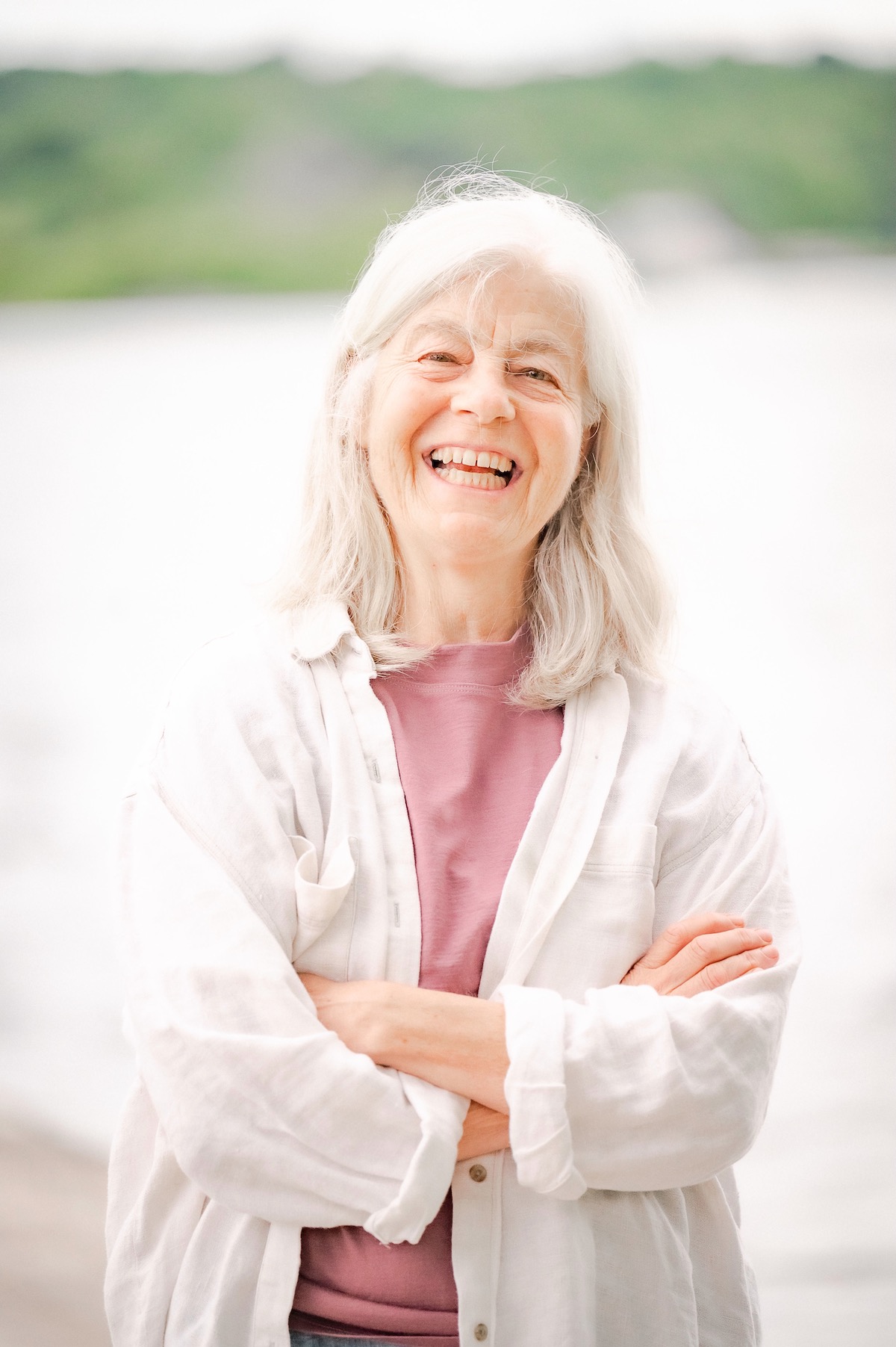 A photo of Susan Wismer. She is a light skin toned woman with mid-length grey hair worn loose, crossing her arms wearing a white linen shirt unbuttoned over a mauve tshirt. She smiles widely. In the background is a lake, with green shores far off on the horizon.