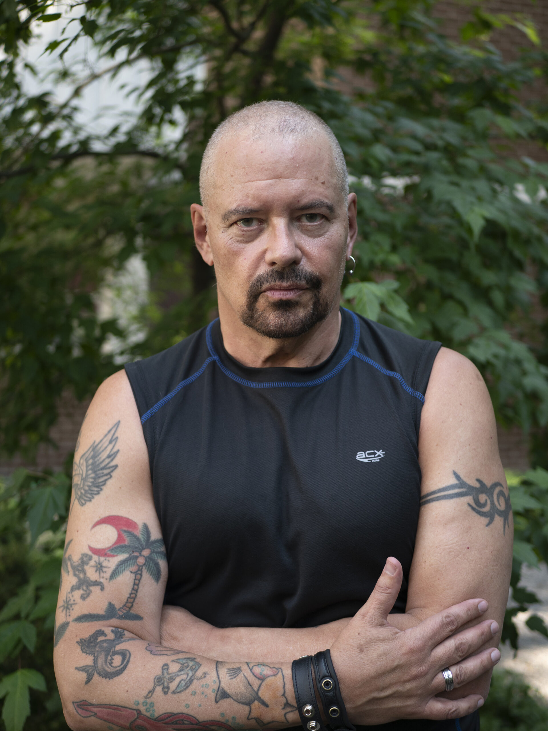 A photo of Sky Gilbert. He is a light-skin-toned man with a shaved head and goatee. He has tattoos on his arms and an earring. He is wearing a black tank top and is standing with his arms crossed in a wooded area.