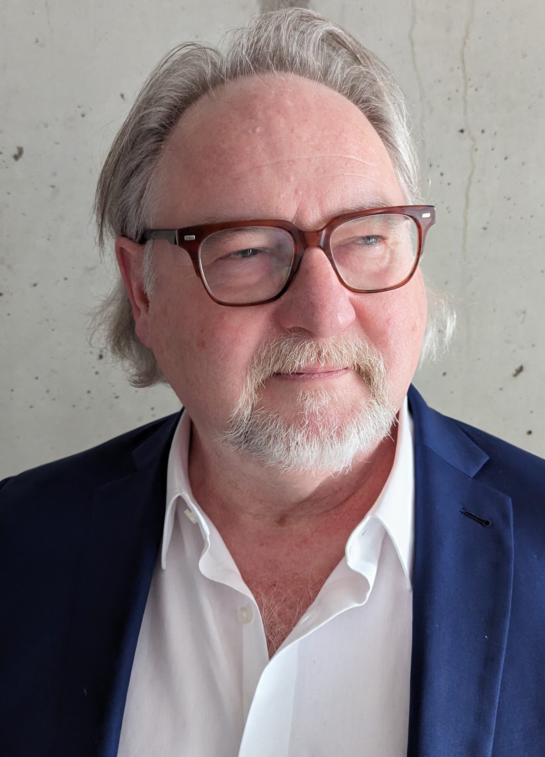 A photo of Vern Thiessen. He is a light skin-toned man with grey hair pushed back and a grey mustache and goatee. He wears brown plastic-framed glasses and a blue suit jacket over a white button-down shirt.