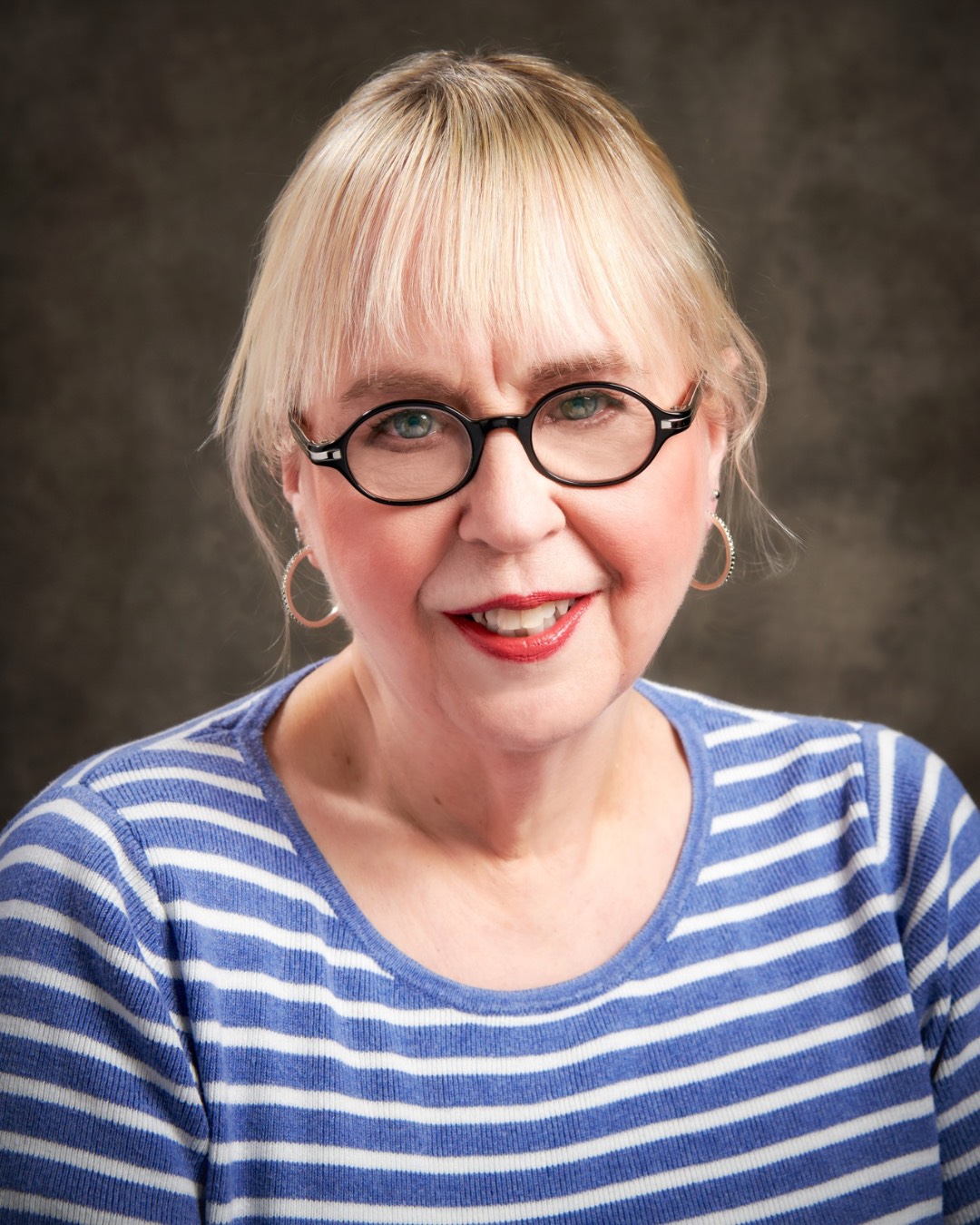 A photo of writer Alice Fitzpatrick. She is a light skin-toned woman with straight blonde hair cut into blunt bangs, worn up. She wears oval-shaped black glasses and gold hoop earrings, and red lipstick. She has a striped blue and white shirt on.