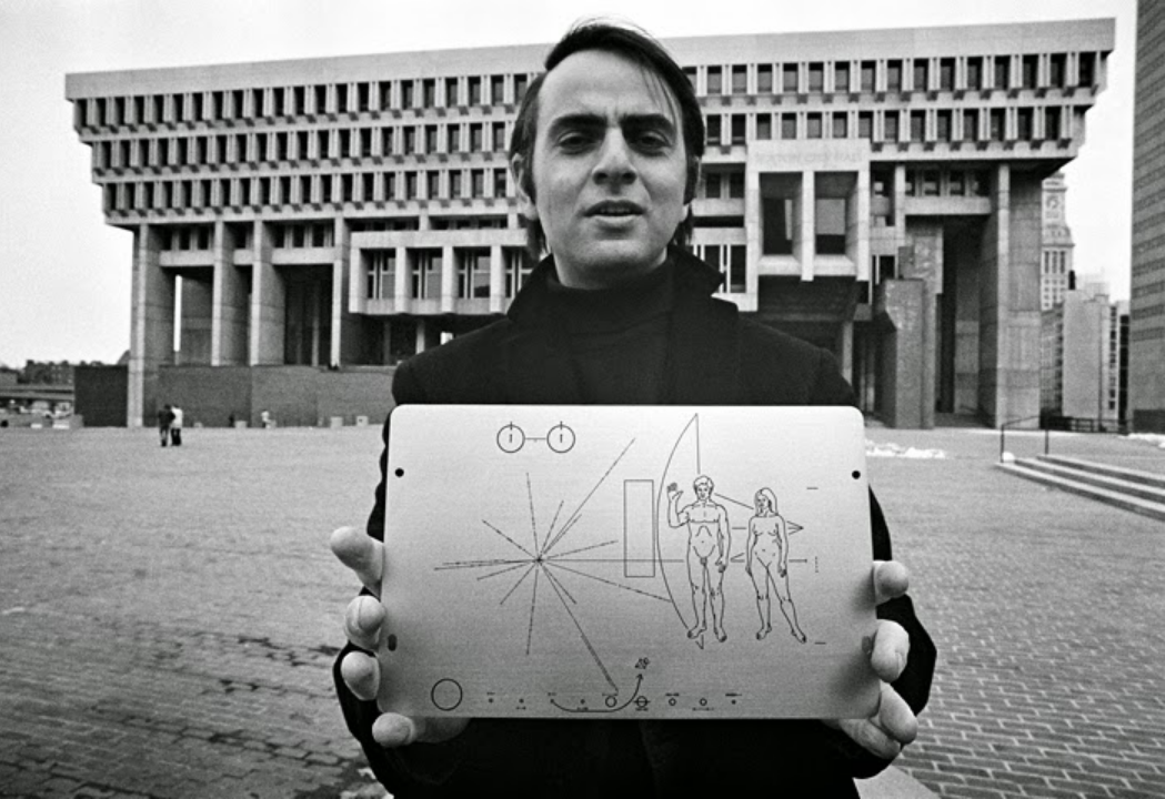 A black-and-white photo of Carl Sagan, a light-skin-toned man with dark, side parted hair, wearing a dark turtleneck and jacket, holding up a plaque to the camera. The plaque shows illustrations of a nude man and woman, a diagram representation of earth within the solar system, and a pulsar reference map.