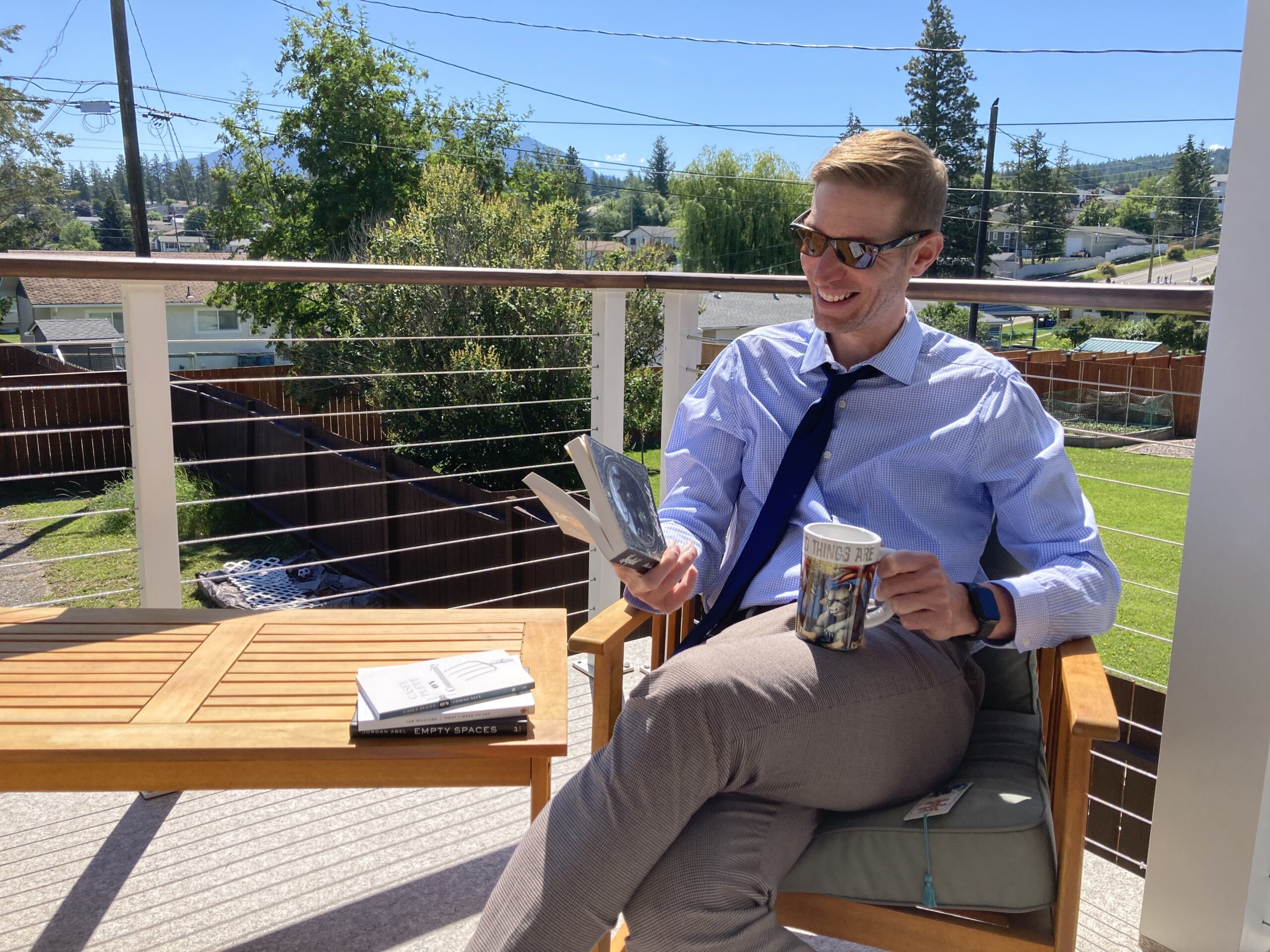 A photo of author Nathan Dueck sitting on a patio on a sunny day. He is a light-skin-toned man with short sandy-blonde hair. He is wearing a black tie, a blue button-up shirt, and khaki pants, and sunglasses. He is smiling while holding a mug and looking at a book.