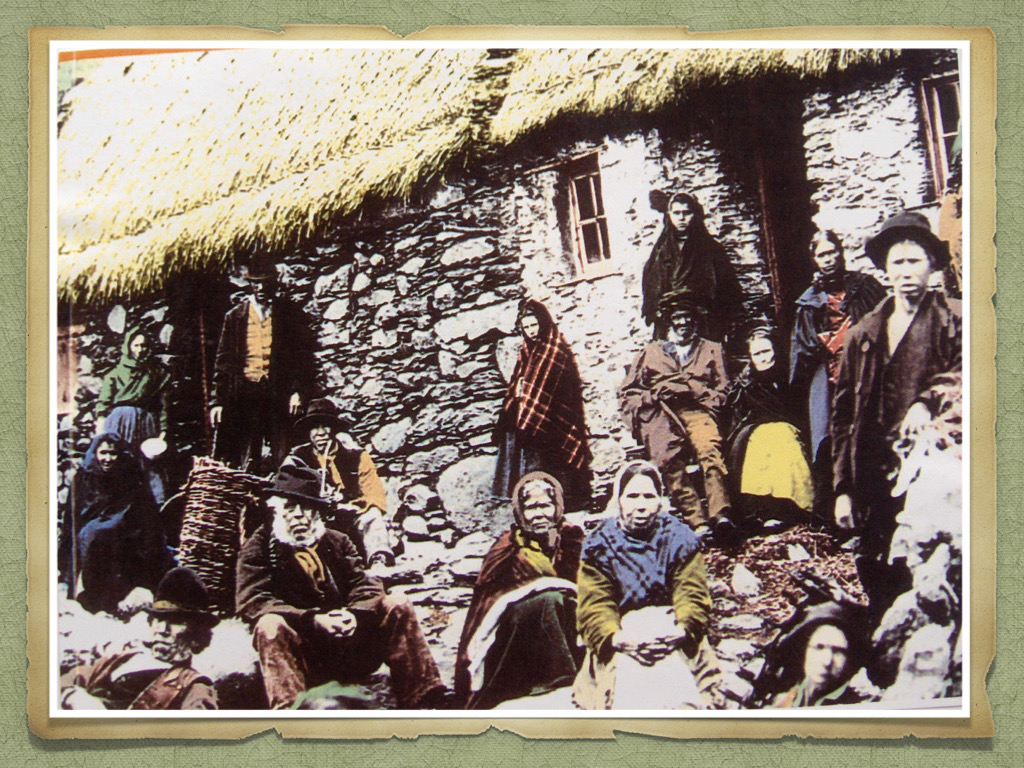 A hand-coloured black and white photo of different people standing and sitting outside of a stone hut with a thatched roof. Among them, a girl with a plaid blanket around her head and shoulders stands apart, staring into the camera.