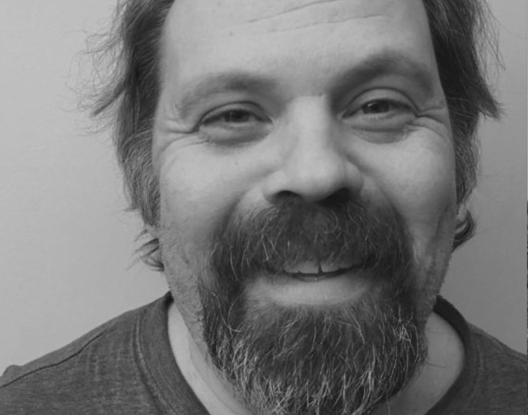 A black-and-white photo of author Shane Neilson. He is a light-skin-toned man with facial hair. He is smiling into the camera. 