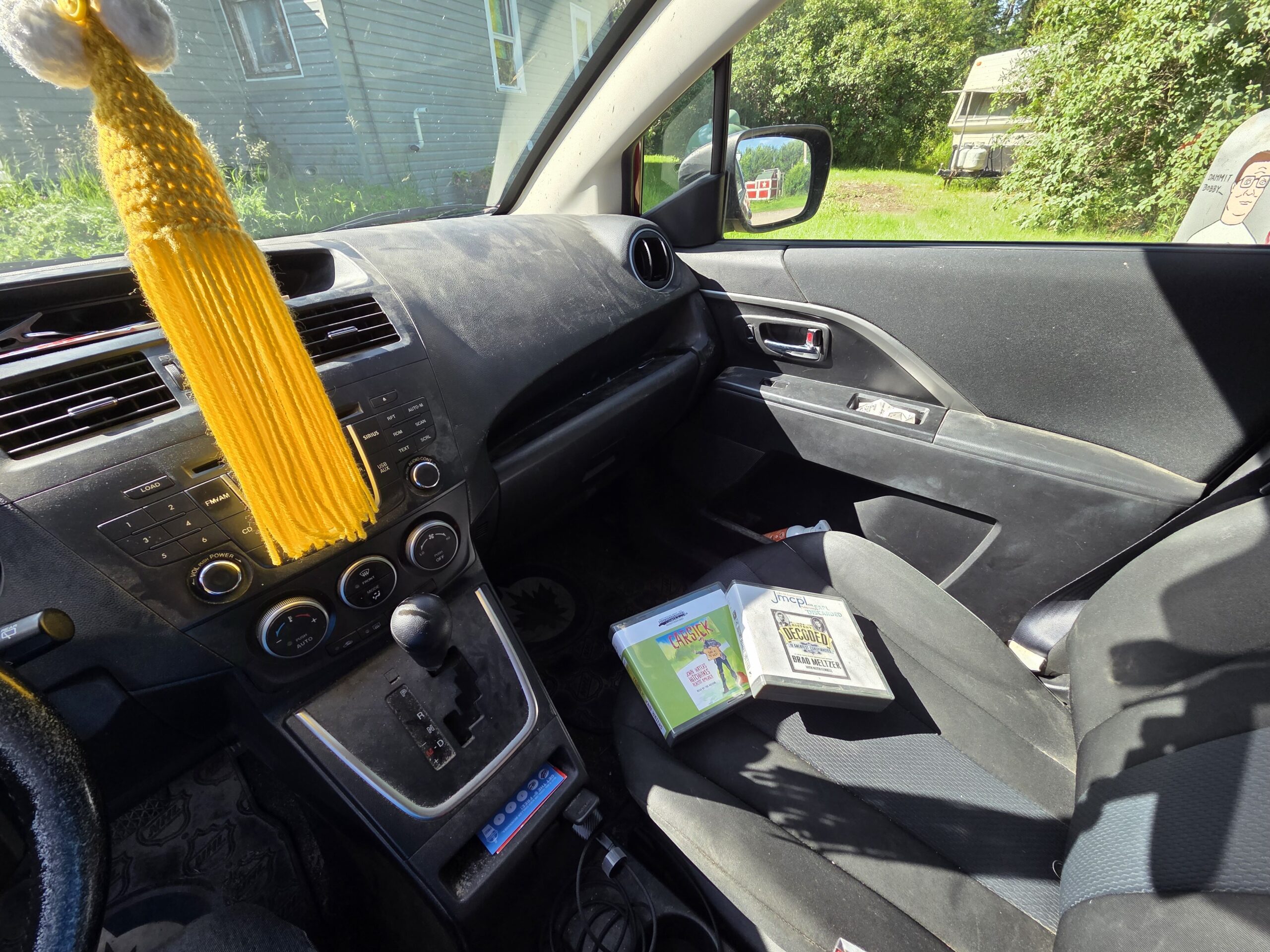 A photo of the interior of author John Brady McDonald's car with a yellow tassel hanging from the rearview mirror, two books resting on the passenger seat, and sunlight streaming through the window.