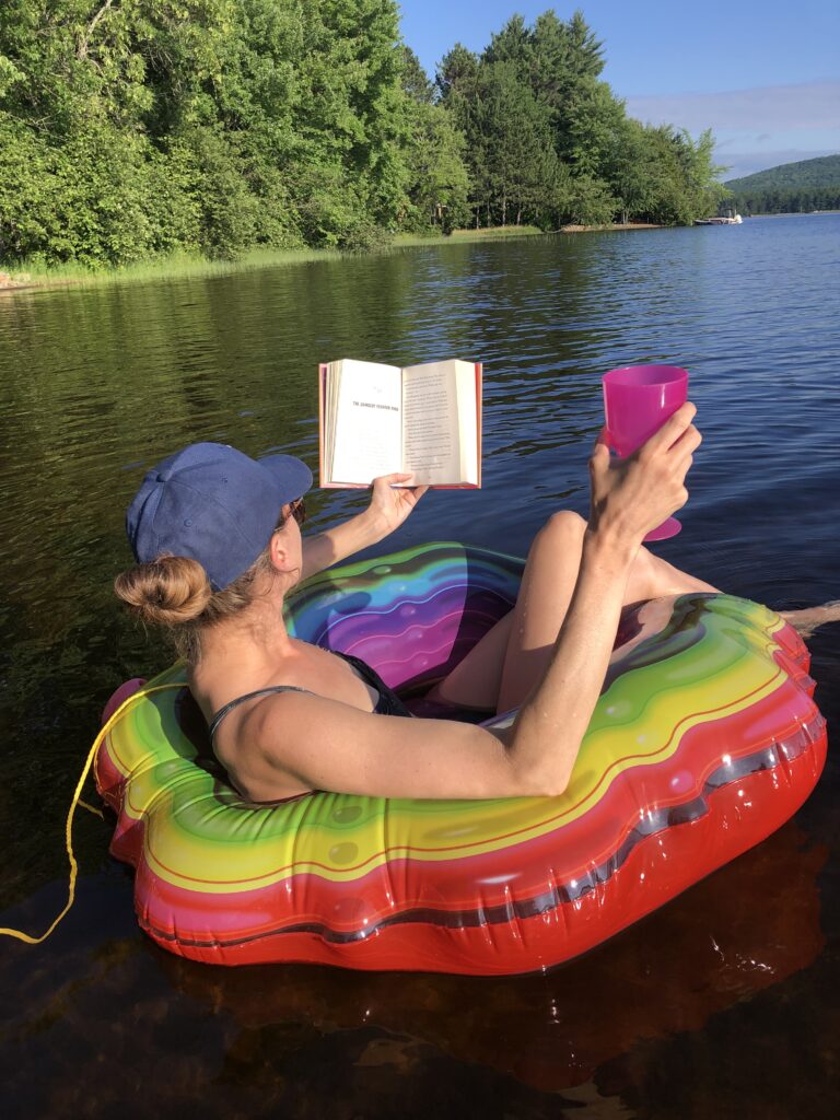 A photo of Lindsay Zier-Vogel from the back lounging on an inflatable water ring with a pink cup in one hand and a book in the other. She is wearing a blue hat and has her hair tied up. 
