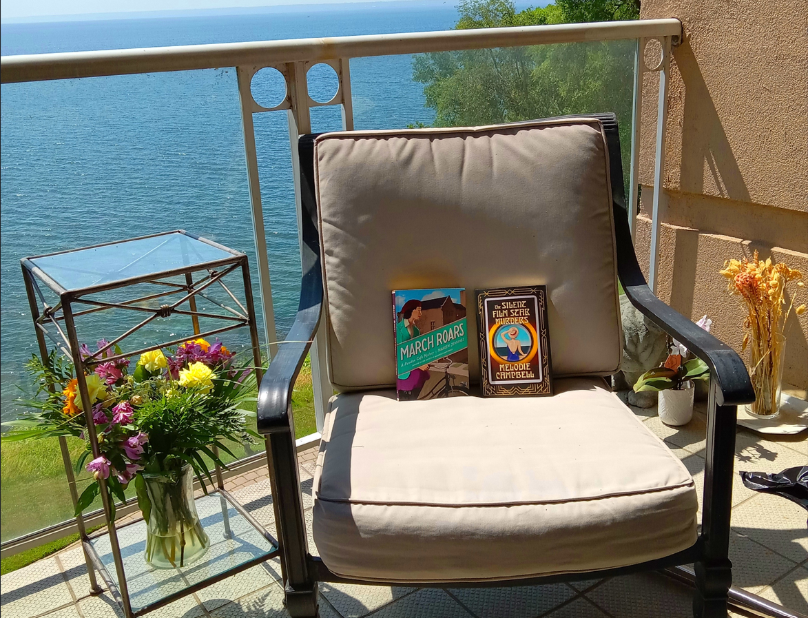 A photo of a balcony with a patio chair with two books on it and table with flowers on it. In the background is a view of the lake and trees. 