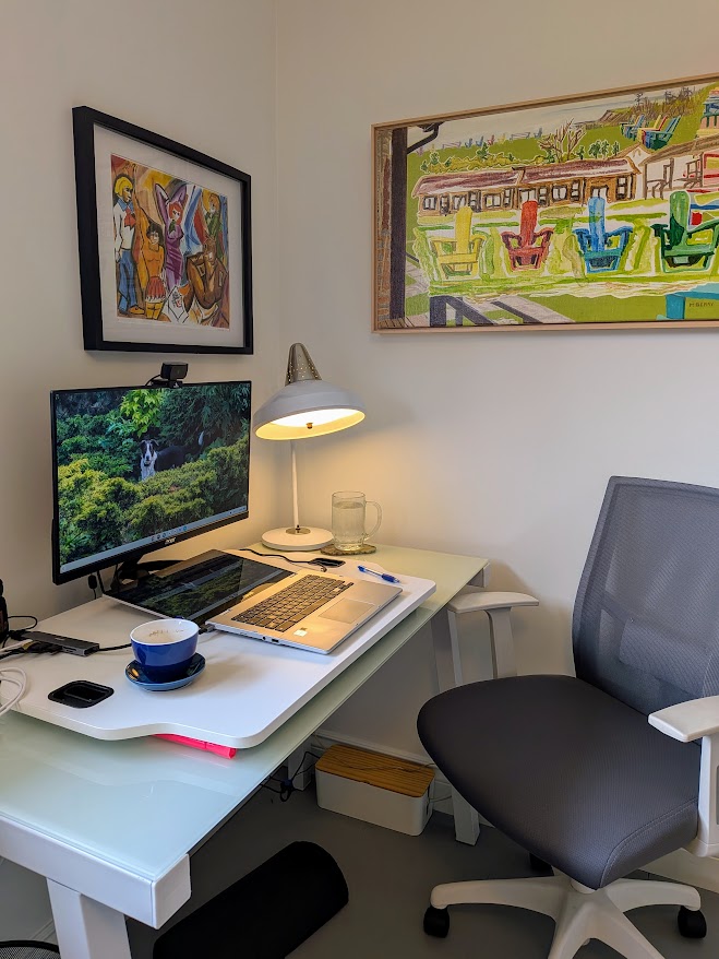 A photo of M.S. Berry's workspace, showing a computer on a desk with a grey chair in front of it, a blue teacup, and a print that looks like a Picasso but it's the group from Scooby-Doo. There is another painting of adirondack chairs on the other wall.