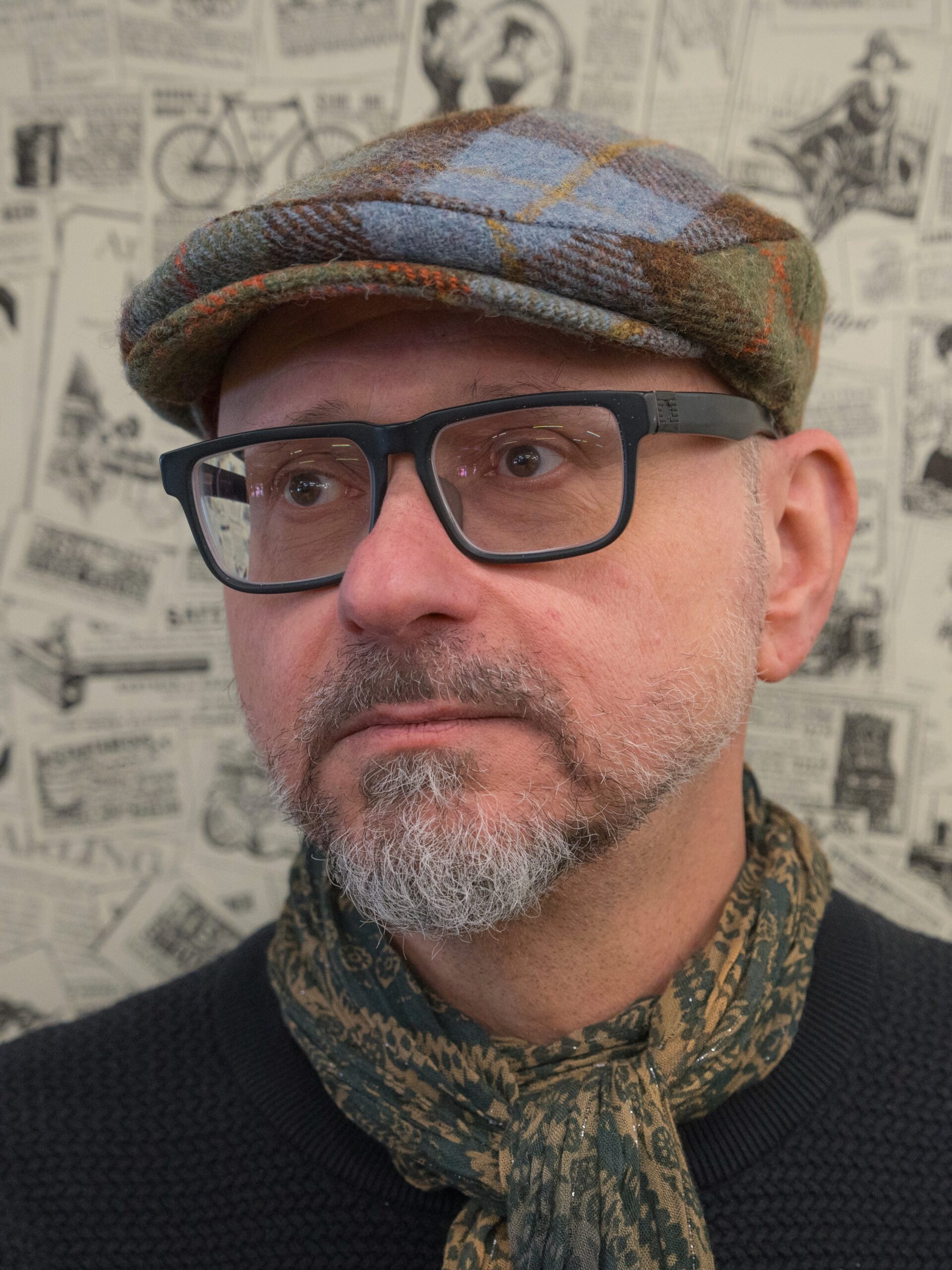 A photo of Richard-Yves Sitoski. He is a light-skin-toned man with greying facial hair. He wears a patterned scarf, a plaid newsboy cap, and dark-rimmed glasses. He is looking off into the distance. 
