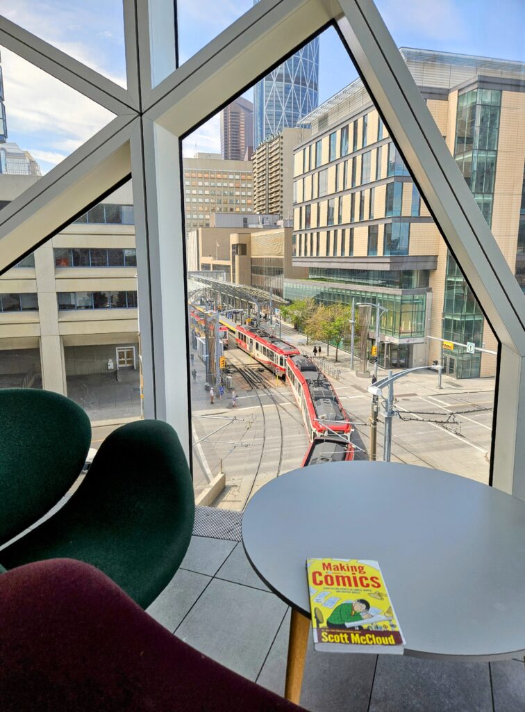 A view from inside the Calgary Central Library overlooking downtown Calgary, with a red C-Train passing below. In the foreground, a round table holds the book Making Comics by Scott McCloud.