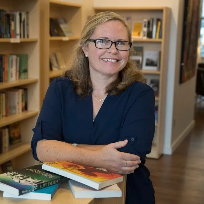A photo of writer M.S. Berry. She is a light skin-toned woman with shoulder-length blonde hair, wearing glasses and a blue button down shirt. She is smiling, crossing her arms and leaning over a pile of books, with bookshelves in the background.