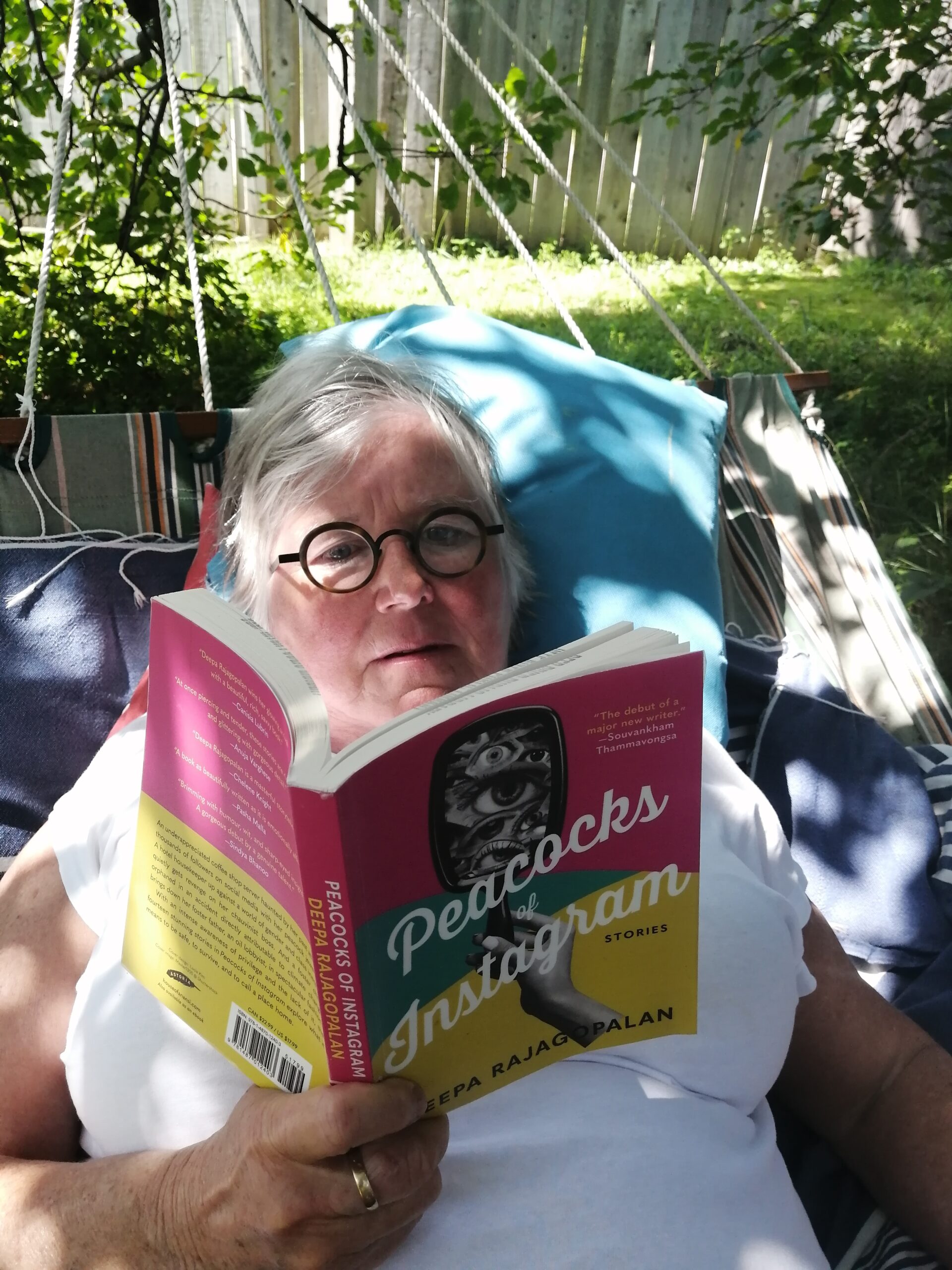 A photo of author Margaret Macpherson laying down on a hammock reading a copy of Peacocks of Instagram by Deepa Rajagopalan. She is a light-skin-toned woman with grey hair and circular black glasses. 
