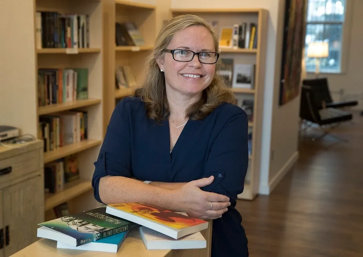 A photo of writer M.S. Berry. She is a light skin-toned woman with shoulder-length blonde hair, wearing glasses and a blue button down shirt. She is smiling, crossing her arms and leaning over a pile of books, with bookshelves in the background.
