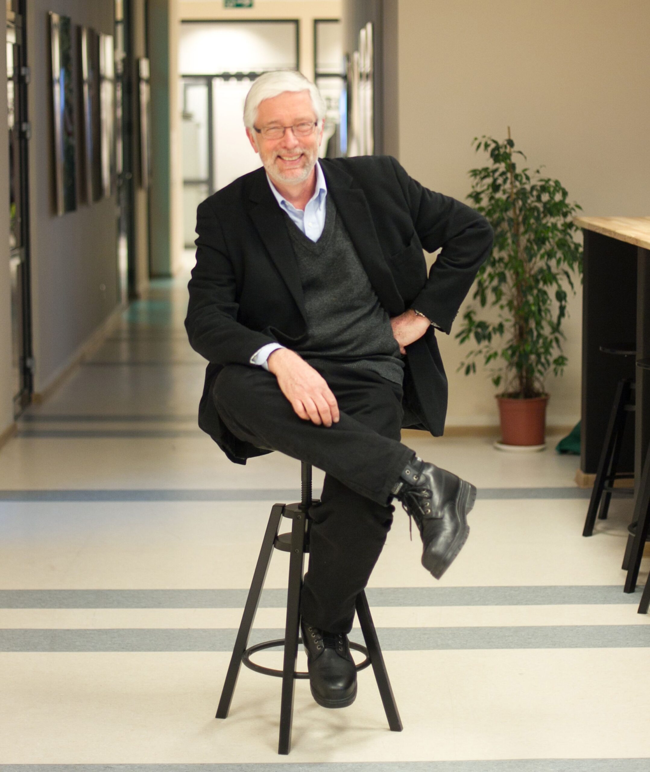 A photo of Antanas Sileika. He is a light-skin-toned man with short white hair parted to the side and a goatee. He is wearing a glasses, and a black suit jacket, black slacks, and a dark grey vest underneath. He is sitting on a stool and smiling into the camera. 