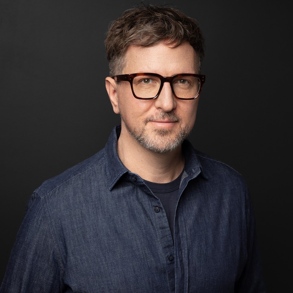 A photo of Bradley Somer. He is a light-skin-toned man with short brown hair and facial hair. He is wearing brown tortoiseshell glasses and a navy-blue collared shirt and smiling into the camera.