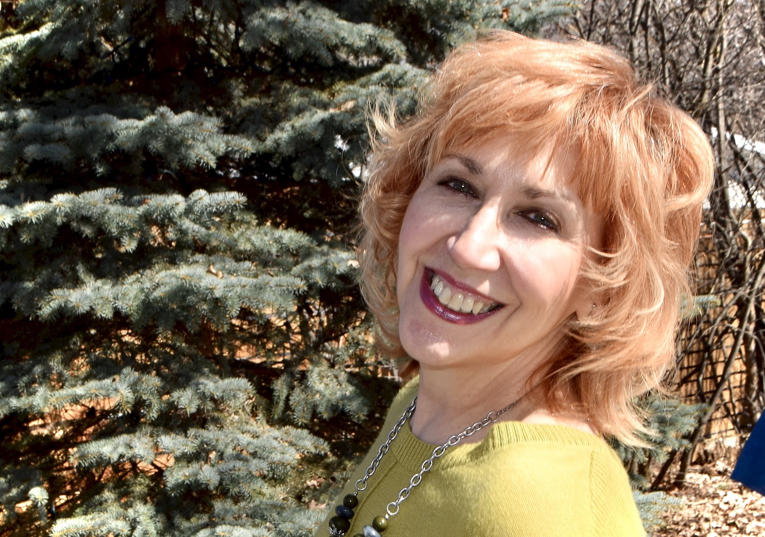 A photo of writer Gail Sidonie Sobat. She is a light skin toned woman with short, brightly coloured orange hair, wearing dark lipstick and a chartreuse sweater, standing beside a pine tree. She smiles widely, squinting into the sunlight.
