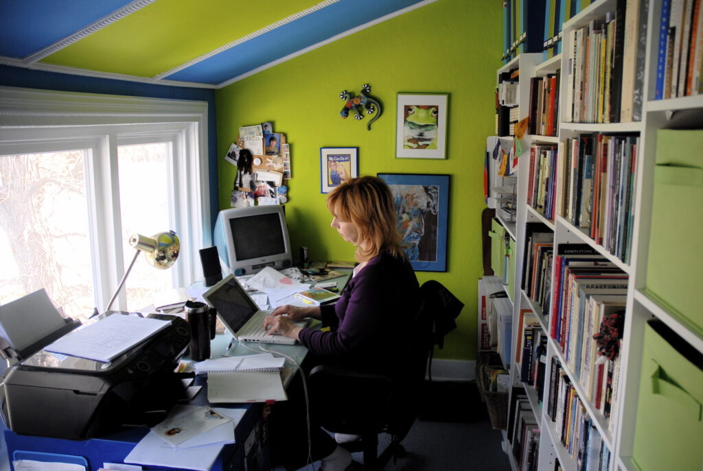 A photo of author Gail Sidonie Sobat in her office. It is a top-floor room with a slanted ceiling, painted in alternating colours of lime green and bright blue. Gail sits and types on a laptop on a crowded desk overlooking a window. There is art on the walls, and loaded white bookcases behind her.