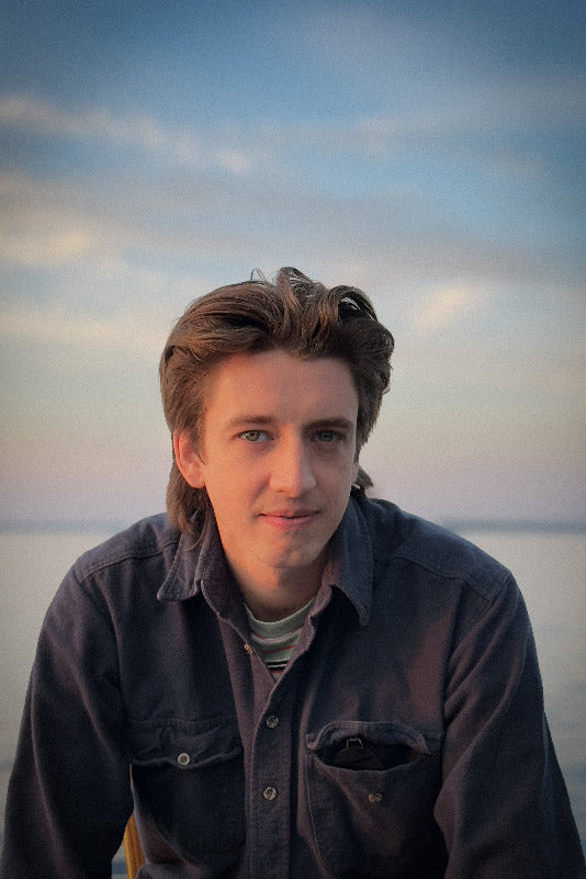 A photo of Guy Elston. He is a light-skin-toned man with light-brown hair. He is wearing a dark-coloured button-up shirt and sitting in front of a body of water. 