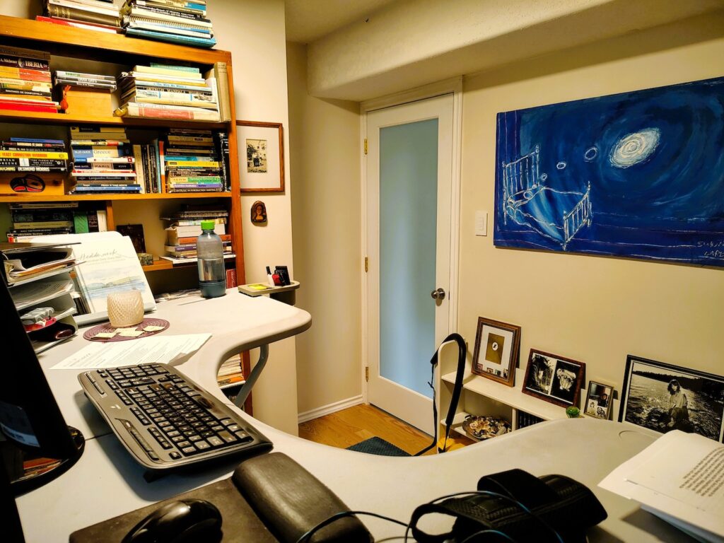 A photo of Marilyn Bowering's office. In the foreground, there is her keyboard and mouse, and the edge of a monitor. A bookshelf laden with books is against the wall. There is a blue painting on the back wall, next to a closed door set in with frosted glass.