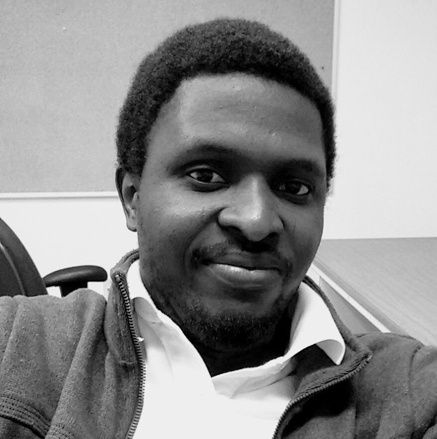A black-and-white close-up photo of James Yékú. He is a black man with facial hair and short dark hair. He is sitting in a chair and smiling into the camera. 