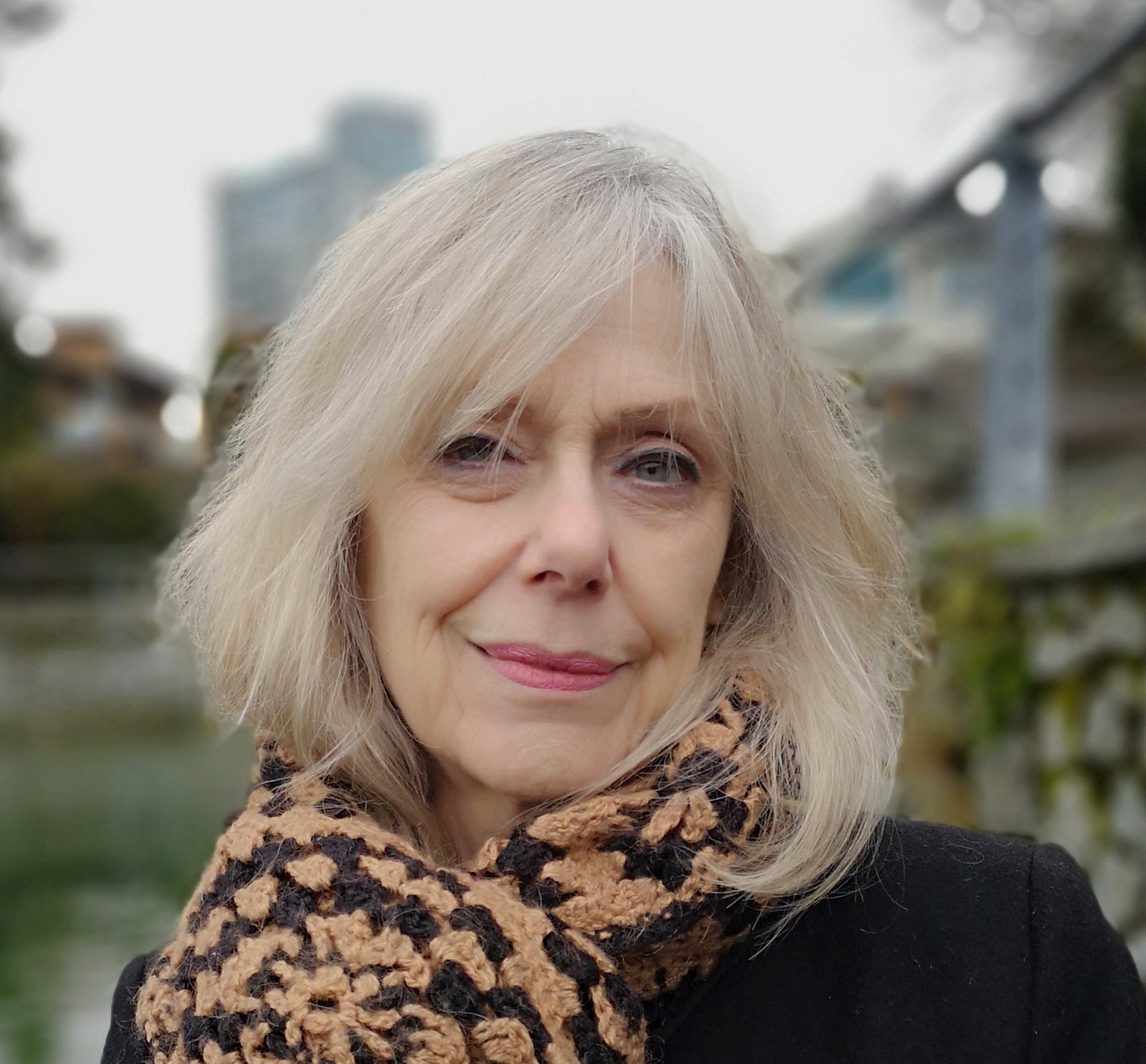 A photo of writer Marilyn Bowering. She is a light skin toned woman with grey/white hair cut into a stylish bob, standing outside, wearing a black coat and crochet scarf. 