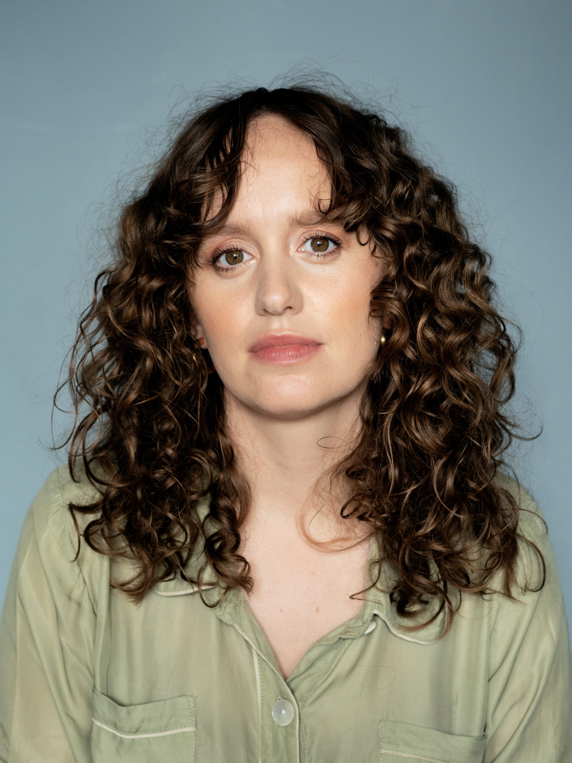 A photo of Olga Ravn. She is a light-skin-toned woman with curly brown hair and brown eyes. She is wearing a light green shirt and looking into the camera.