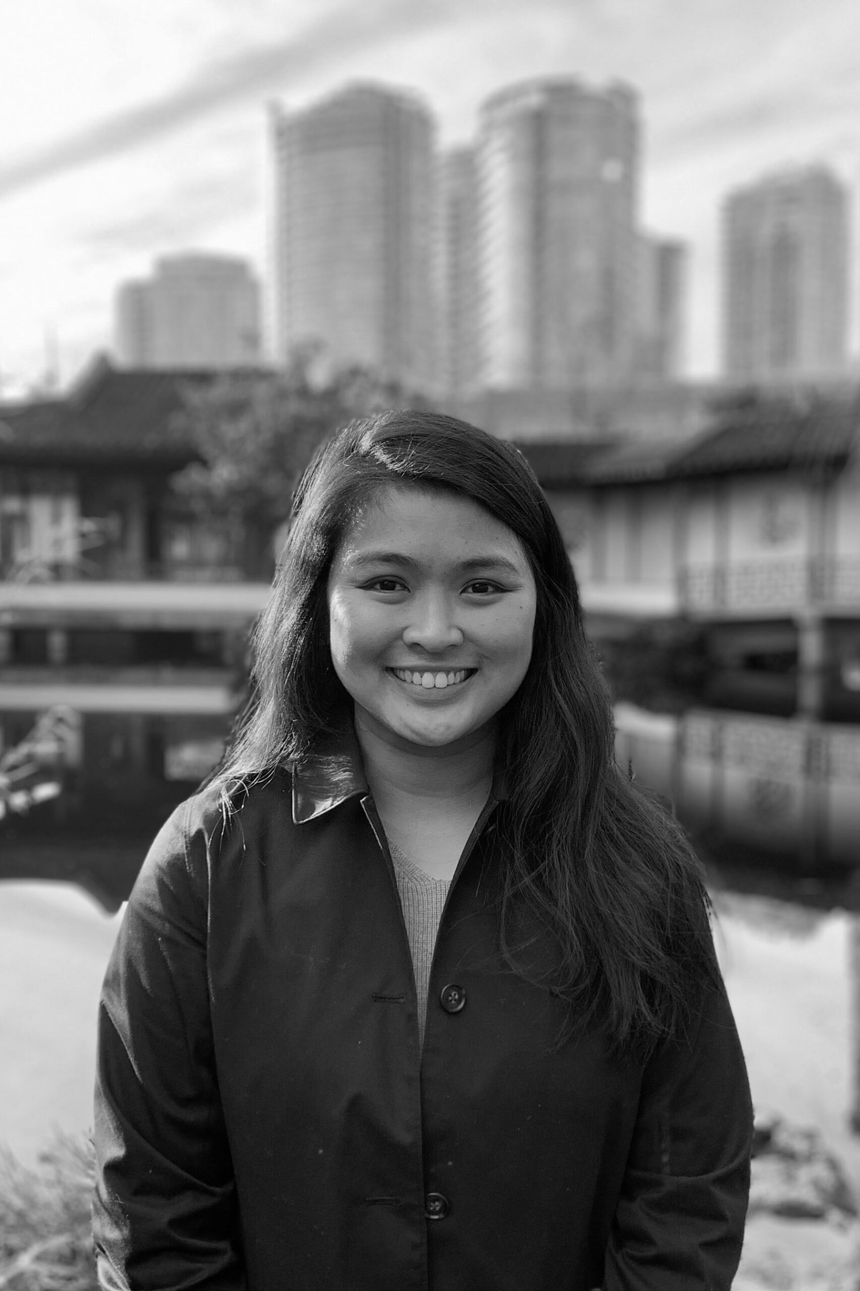A black-and-white photo of Steffi Tad-y. She is a woman of filipino descent with long brown hair and brown eyes. She is wearing a black coat with a collar and standing with the cityscape behind her and smiling into the camera.