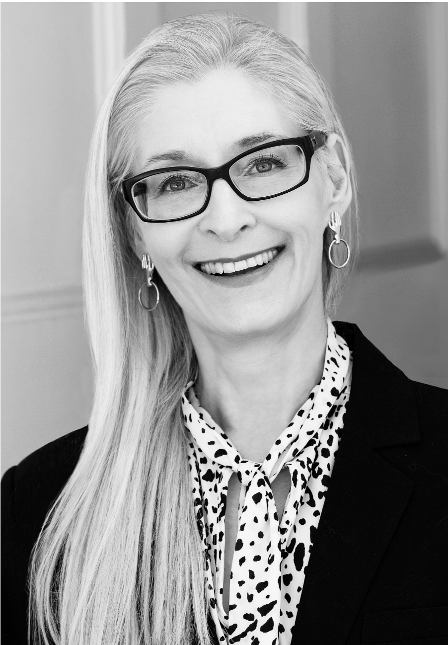 A black and white photo of writer Lisa de Nikolits. She is a light skin-toned woman with long, straight blonde hair and glasses, wearing a spotted blouse and dark blazer. She smiles widely.