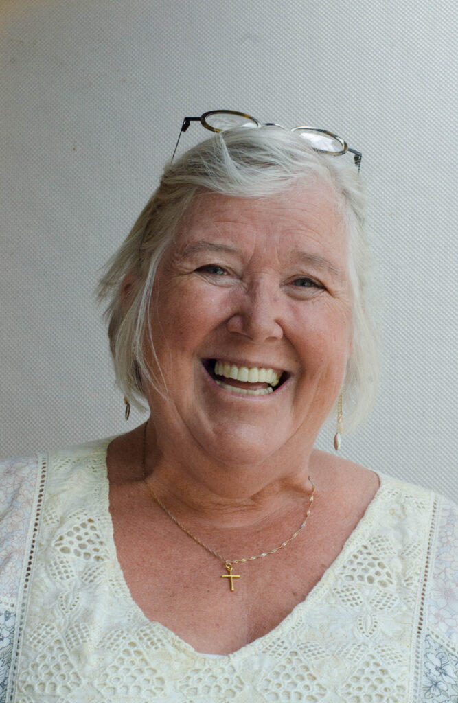 A photo of Margaret Macpherson. She is a light-skin-toned woman with short grey hair. She is wearing a white top, a gold necklace with a cross, and glasses on top of her head. She is standing in front of a grey background and smiles into the camera. 