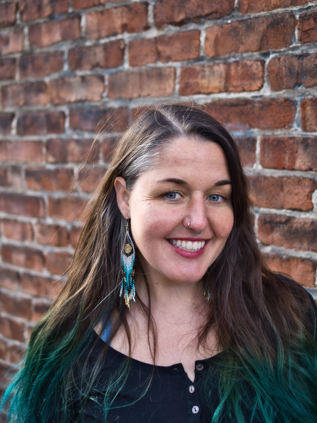 A photo of Alex D. Ketchum. She is a light-skin-toned woman with long brown hair with green tips. She is wearing a dark-blue shirt, dangly earrings, and smiling into the camera in front of a brick wall.