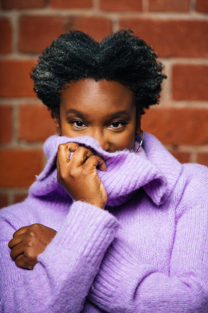 A photo of Andrea Scott. She is a Black woman with short curly hair, wearing a lilac-coloured turtleneck. She is standing in front of a brick wall and hiding part of her face in the cowl of her turtleneck with her arms crossed. She appears to be smiling. 