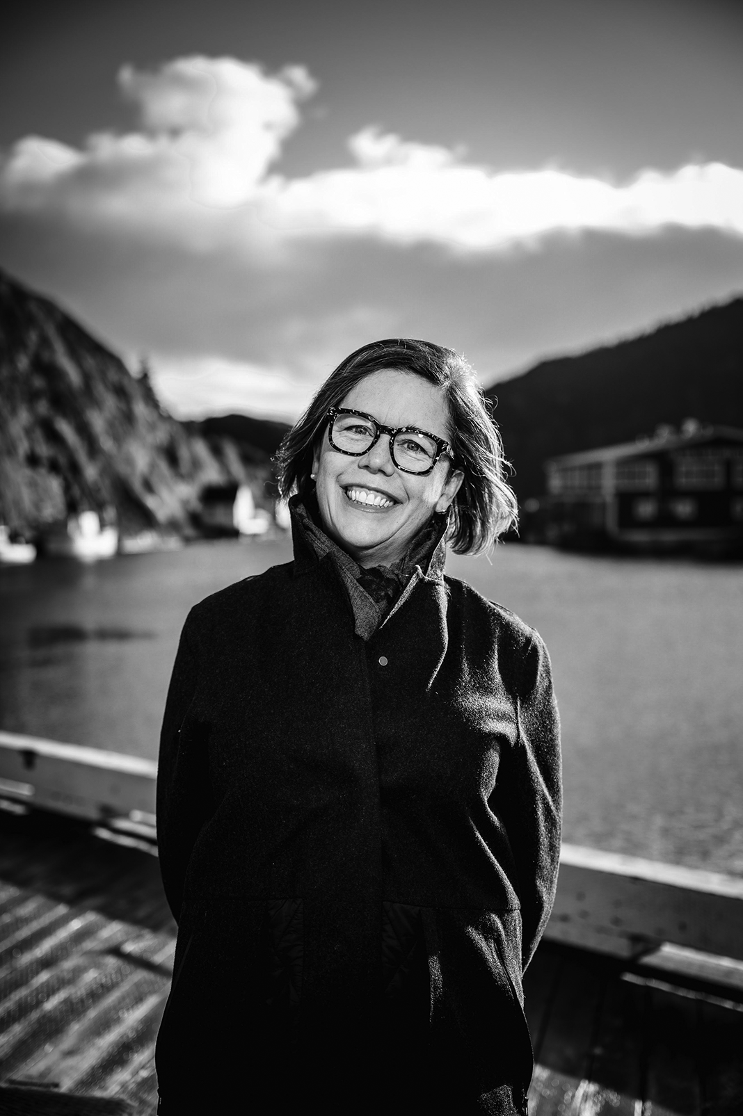 A black-and-white photo of Angela Antle. She is a light-skin-toned woman with chin-length hair. She is standing outdoors on a wooden dock with water and hills in the background. She is wearing glasses, a dark coat, and a scarf, with sunlight illuminating her face and clouds visible in the sky behind her.