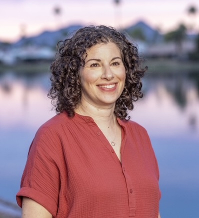 A photo of Claire Sicherman. She is a light-skin-toned woman with curly brown hair. She is wearing a coral-coloured button-up shirt and standing near a body of water. 