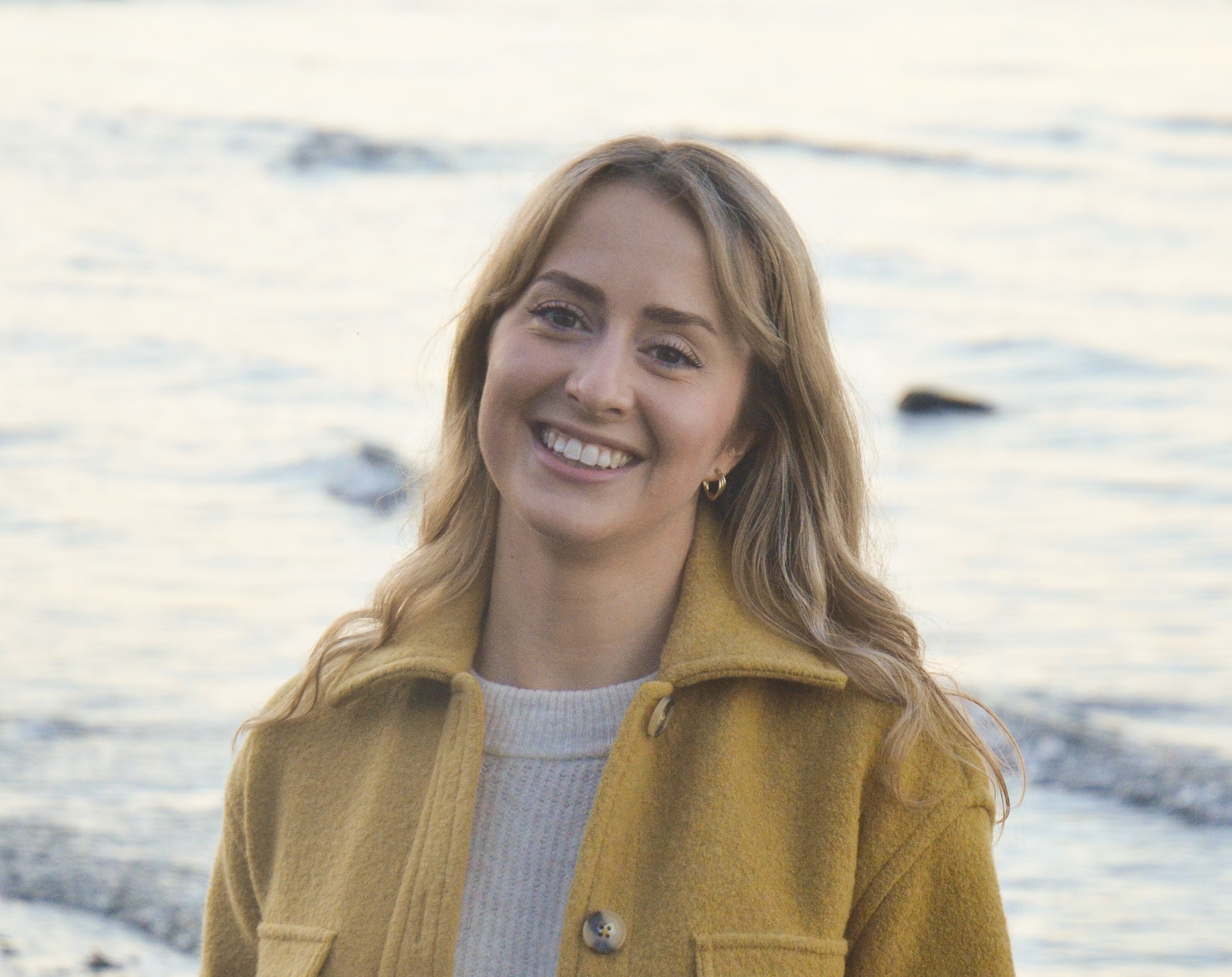 A photo of Jazmin Welch. She is a light-skin-toned woman with shoulder-length blonde hair. She is wearing a yellow jacket and standing in front of a body of water smiling into the camera. 