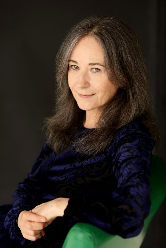 A photo of Elise Levine. She is a light-skin-toned woman with light brown hair past her shoulders. She is sitting on a green chair with her arm draped on one side and her body angled slightly away from the camera. 