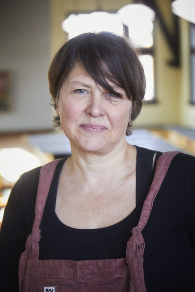 A photo of Heather Birrell. She is a light-skin-toned woman with short brown hair. She is wearing a black top and rose-coloured corduroy overalls and standing indoors in soft natural light.